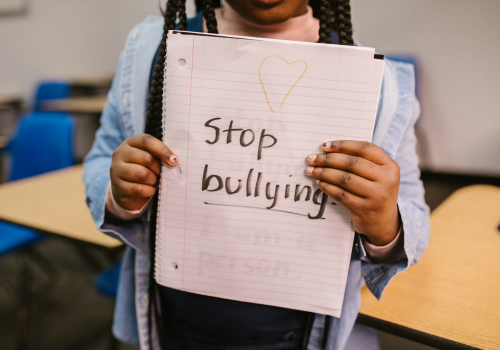 Photo of a young child from the chin down in a classroom holding a handwritten sign saying "Stop bullying" 