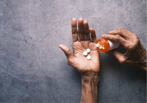 Photo of a pair of skinny, elderly hands, one holding several pills & the other holding the pill bottle