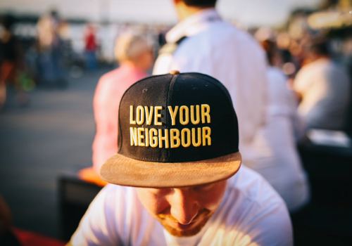 Photo of a man in a white t-shirt wearing a black and tan hat saying "Love Your Neighbor." He's sitting in a crowd.