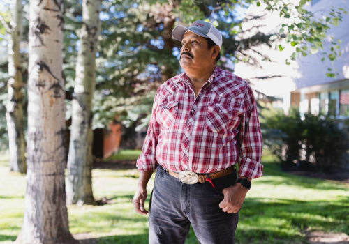 An Indigenous man wearing a red plaid shirt, black jeans, boots and ball cap