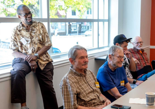 Photo of a Black man laughing at a window & 4 white men sitting at a table smiling