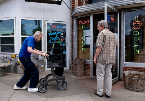 Photo of a frail elder pushing his walker toward the open door held by another elder