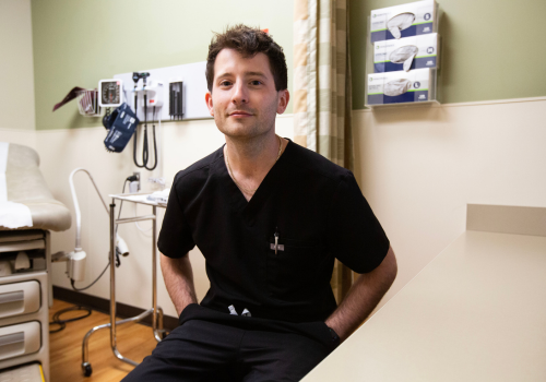 A Gay white man wearing black scrubs sits at an exam table