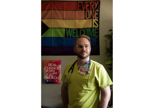 A Gay white man wears yellow scrubs & stands in front of a rainbow banner.