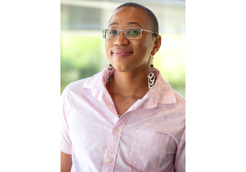 A Black Trans man wears a pink shirt w/white dots, glasses and long earrings