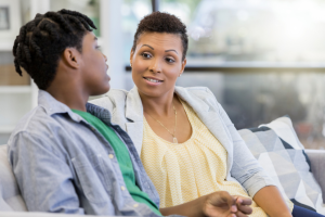 A Black mother and teen son sit on a sofa talking