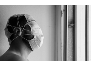 Side view of a man wearing medical masks covering his entire head. He faces a barred window.