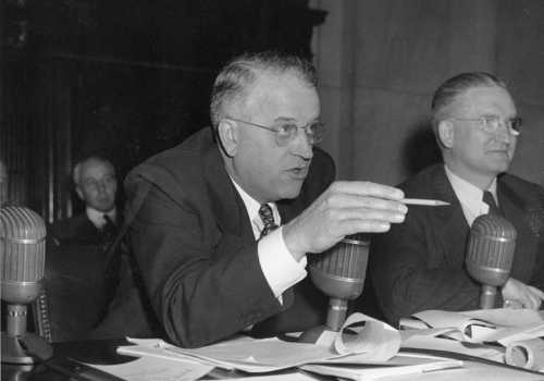 Two white men wearing glasses sit at a table with microphones. Wherry gesticulates with a pencil as he speaks.