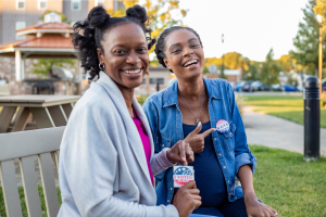 Two, young Black women sit on a park bench smiling & holding "I Voted" stickers