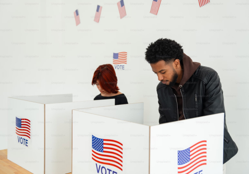 A young, Black man stands in a cubby & votes next to an Asian-looking woman in her own cubby