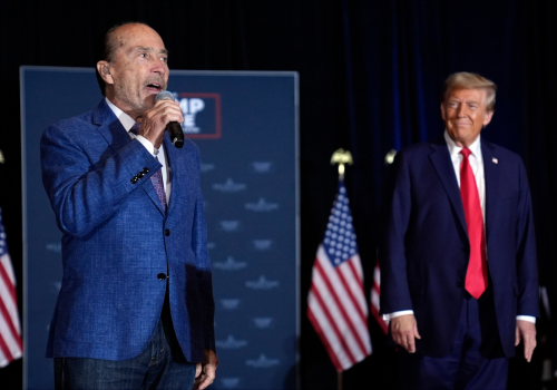 One balding, white man in a blue suit sings as another white man in a suit & red tie listens smiling