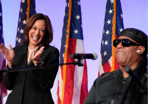 A Black woman in a dark suit claps & smiles as a Black man in sunglasses sings