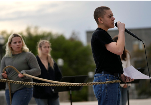 A Trans youth w/cropped hair & wearing a black shirt & jeans speaks into a mic while wrapped w/a rope held by another student