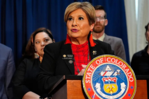 Monica Duran stand behind a lectern with the State of Colorado seal and surrounded by other people.