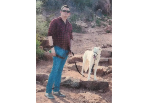 A man in a dark, red shirt & sunglasses stands w/his white dog on some rock steps