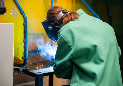 A woman with a red ponytail and wearing a green shirt and welding helmet works in a safety cubicle