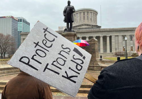 Trans youth advocates holding a "Protect Trans Kids" sign stand in front of a statute