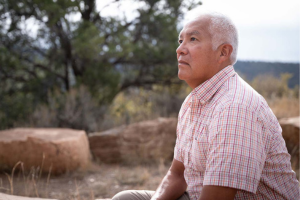 Photo of a middle-aged Indigenous man wearing a short-sleeve shirt & sitting near trees & rocks