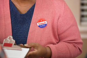 Close-in photo of a Black woman's chest and hand; she wears an "I Voted" pin and is holding a clipboard