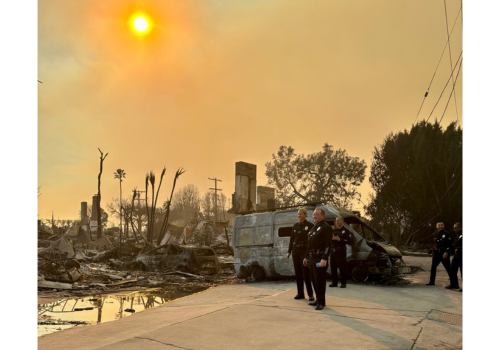 Several officers survey a burned out scene with a smoky sky making the sun orange