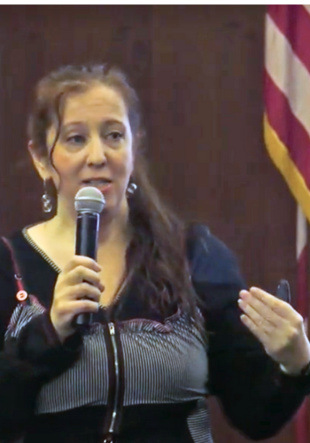 A white woman with long, brown hair holds a microphone and gestures