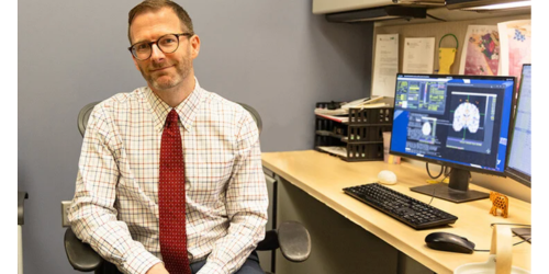 A white man with glasses and facial hair sits as a desk next to screen with a brain image