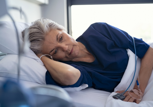 A white woman in a blue top & w/gray hair lies in a hospital bed w/tubes