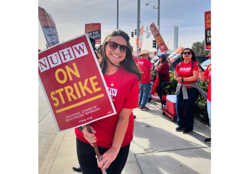 A woman wearing a red shirt & holding a sign saying, "NUHW On Strike" among other striking workers